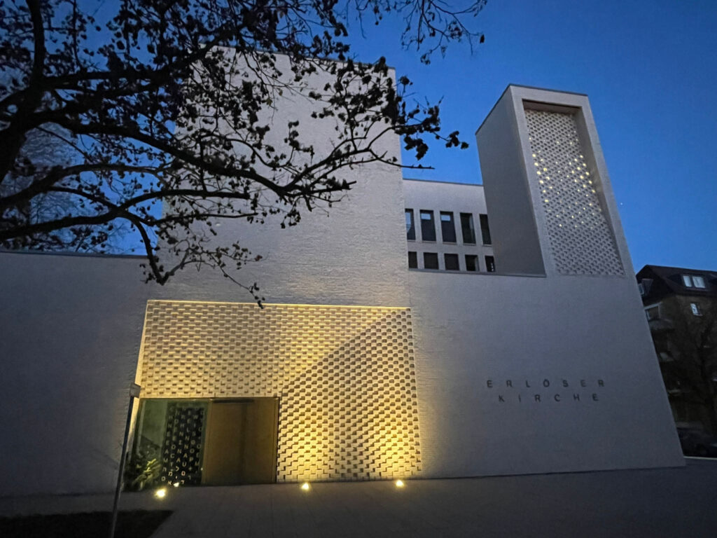 Die neue Erlöserkirche in Köln Weidenpesch mit Ansicht auf das Hauptportal und den Glockenturm. Das Portal wird im Abendlich hell angestrahlt - der Glockenturm ist von innen beleuchtet und schimmert gelblich. Im oberen rechten Bereich wird das Kirchengebäude von einem herblichen Baum mit Ästen verdeckt_Foto Manfred Buge