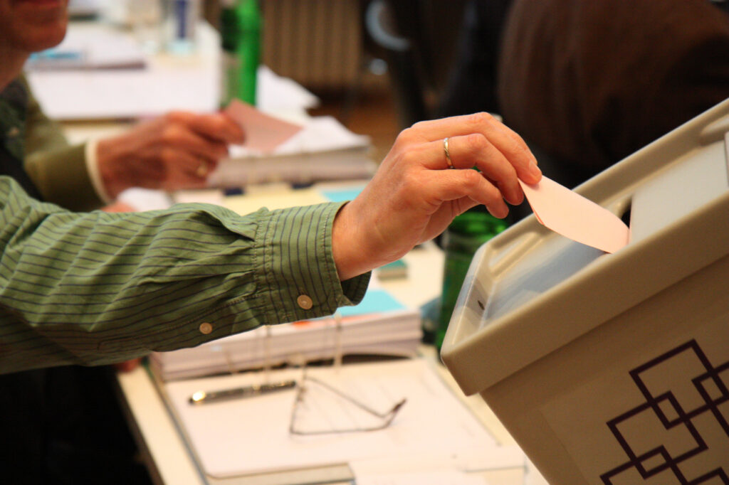 Ein Stimmzettel der Wahl wird in eine kirchliche Wahlurne eingeworfen - der Stimmzettel ragt noch zur Hälfte heraus und wird von einer Hand festgehalten
