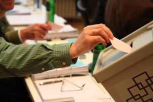 Ein Stimmzettel der Wahl wird in eine kirchliche Wahlurne eingeworfen - der Stimmzettel ragt noch zur Hälfte heraus und wird von einer Hand festgehalten