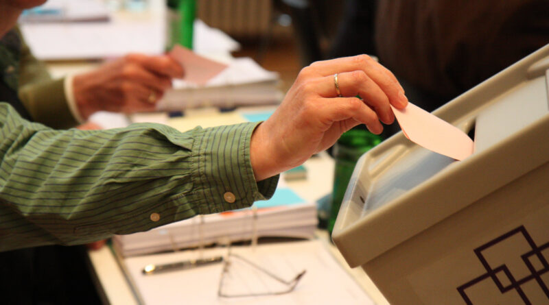 Ein Stimmzettel der Wahl wird in eine kirchliche Wahlurne eingeworfen - der Stimmzettel ragt noch zur Hälfte heraus und wird von einer Hand festgehalten